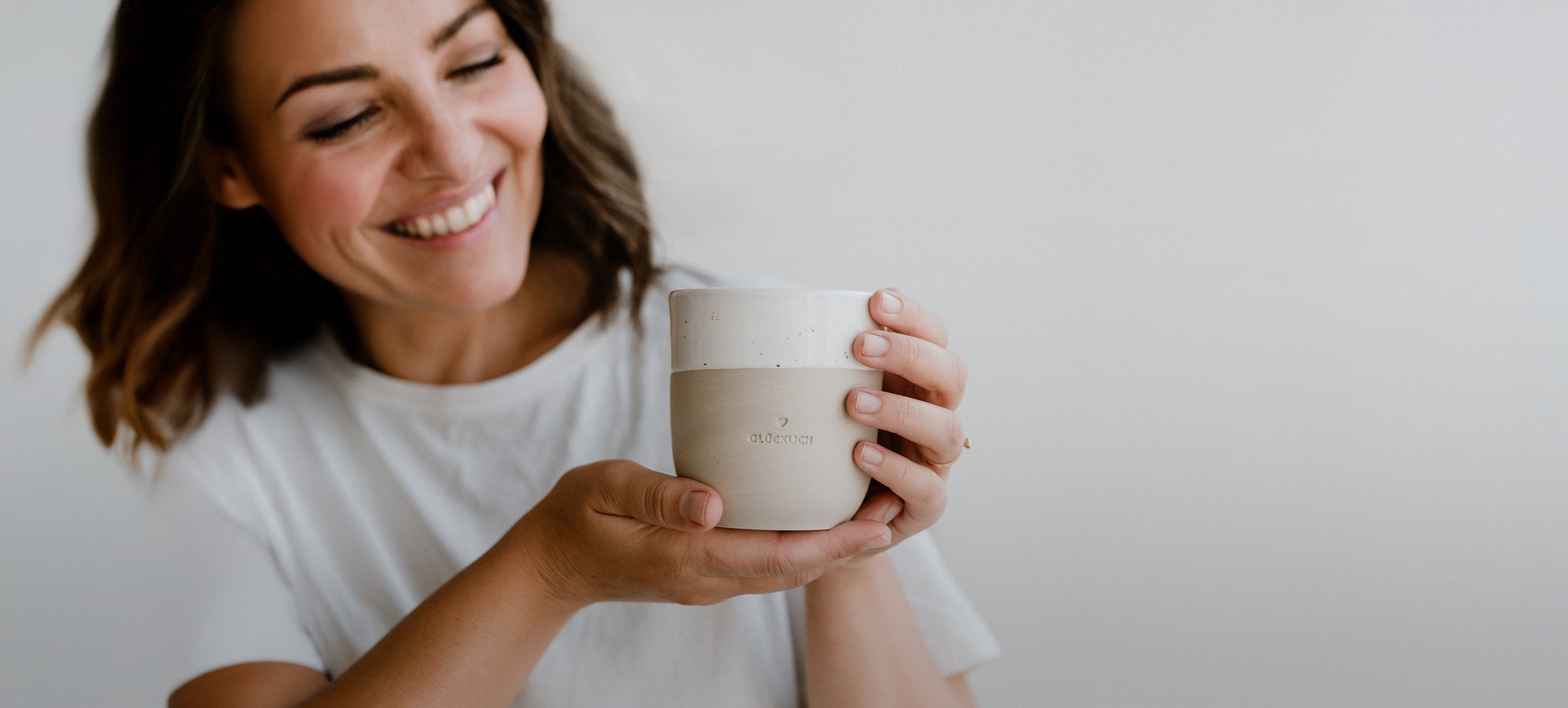 Lächelnde Frau in weißem T-Shirt hält einen hellen Steingutbecher mit Blindprägung ‚Glücklich‘ in beiden Händen vor sich.
