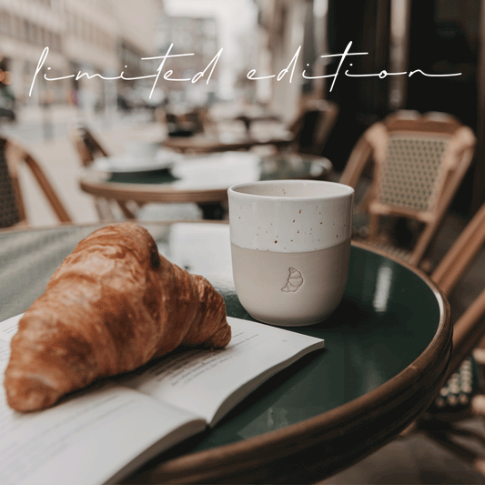 Videosequenz mit verschiedenen Situationen mit dem limitierten Becher "Croissant". In den Händen einer Frau mit einem braunen Wollpullover. Auf einem dunkelgrünen Tisch eines Cafes. Nahaufnahme der Prägung des Bechers.