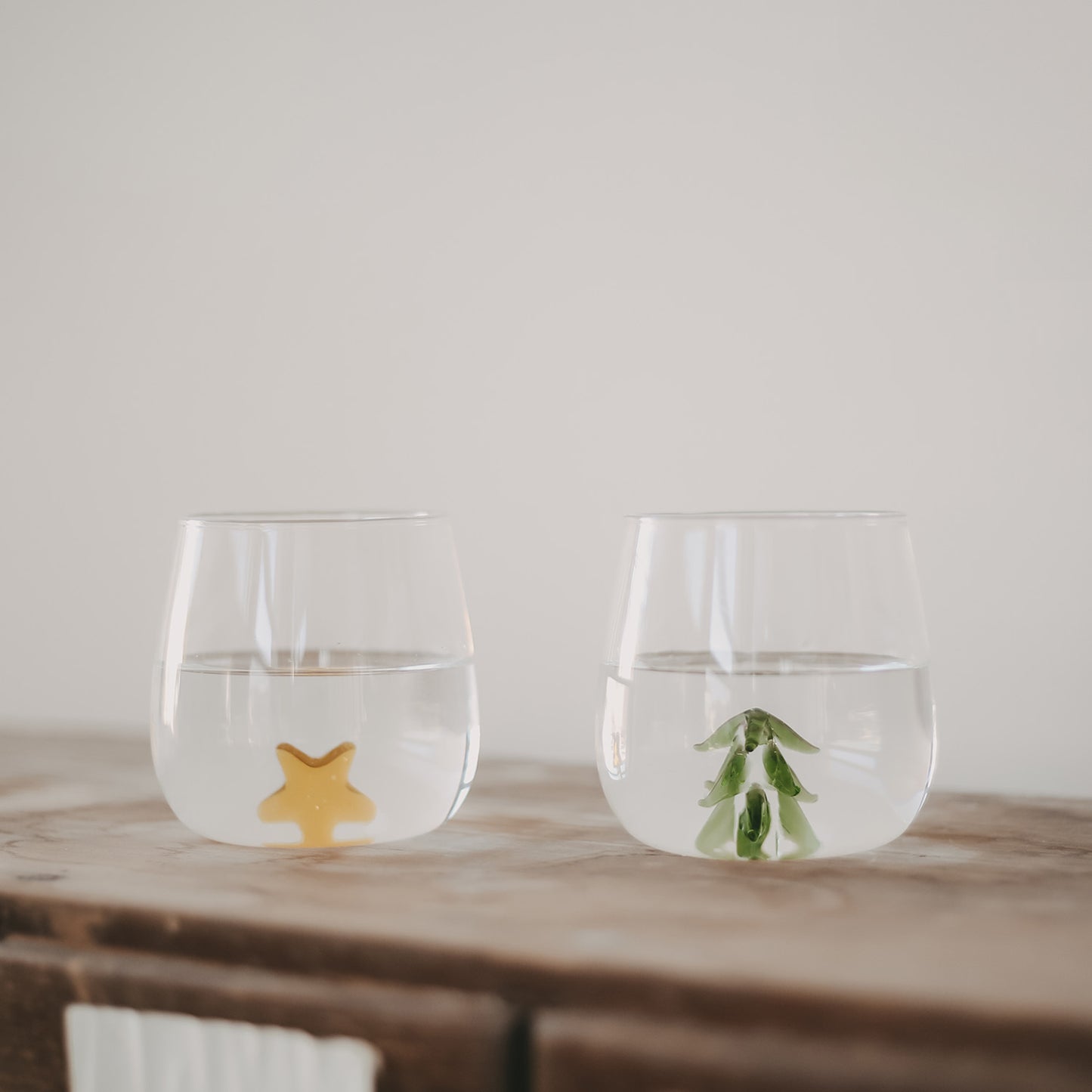 Zwei Trinkgläser mit Wasser, eines mit einer gelben Sternfigur, das andere mit einer grünen Tannenbaumfigur, auf einem Holztisch