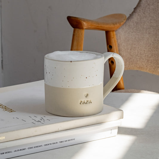 Keramiktasse mit Henkel in Beige und weiß gesprenkelter Glasur, auf einem gestapelten Buchstapel aus Kunst- und Designbüchern platziert. Auf der Vorderseite ist ein kleines Herz und das Wort ‚PAPA‘ eingeprägt. Die Tasse ist mit Milchschaum gefüllt. Im Hintergrund ein minimalistisches Holzstuhl-Detail und eine natürliche Lichtstimmung – liebevoll gestaltet als stilvolles Geschenk für Väter.