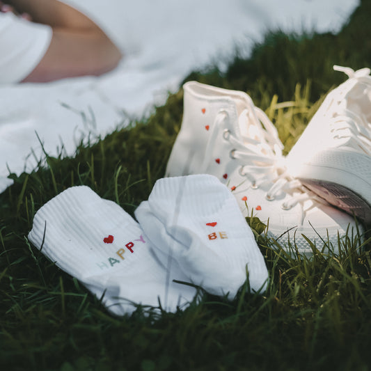 Weiße Socken mit der Aufschrift ‚BE' mit rotem Herzchen auf der einen und 'HAPPY‘ mit rotem Herzchen auf der anderen Ferse. Liegend im Gras neben weißen Sneakern.