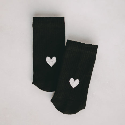Schwarze Socken mit weißen Herz-Motiv, nebeneinander auf hellem Untergrund liegend.