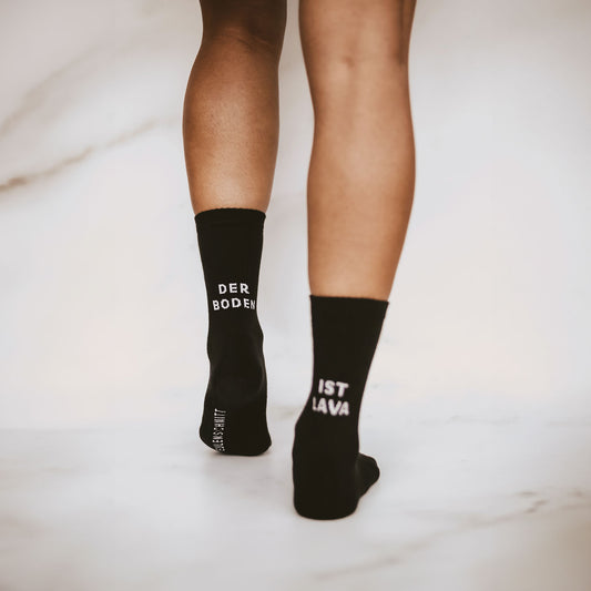 Person von hinten fotografiert, trägt schwarze Socken mit der weißen Aufschrift DER BODEN IST LAVA auf den Fersen. Nur Beine und Socken sind sichtbar.