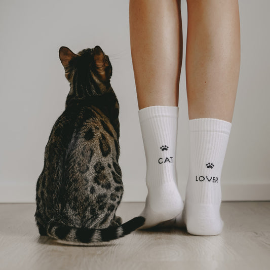 Weiße Socken mit schwarzem Aufdruck „CAT“ und „LOVER“ sowie Pfoten-Symbolen an den Waden einer Frau; daneben sitzt eine getigerte Katze auf einem hellen Boden und blickt nach oben.