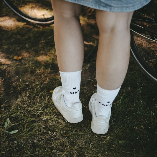 Eine Frau steht auf einer Wiese neben einem Fahrrad. Sie trägt weiße Sneaker und weiße Socken mit schwarzem Text: Auf der linken Socke steht 'CIAO', auf der rechten 'KAKAO', jeweils mit einem kleinen schwarzen Herz darüber.
