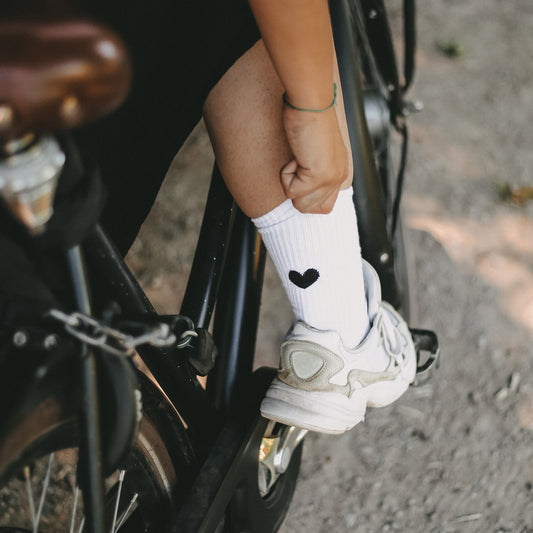 Person sitzt auf einem Fahrrad und zieht eine weiße Socke mit schwarzem Herzmotiv hoch – sportlich-lässige Szene auf einem Schotterweg.