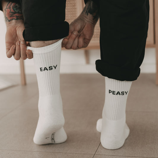 Person mit Tätowierungen trägt weiße Socken mit den Schriftzügen 'EASY' auf der einen und 'PEASY' auf der anderen Ferse.
