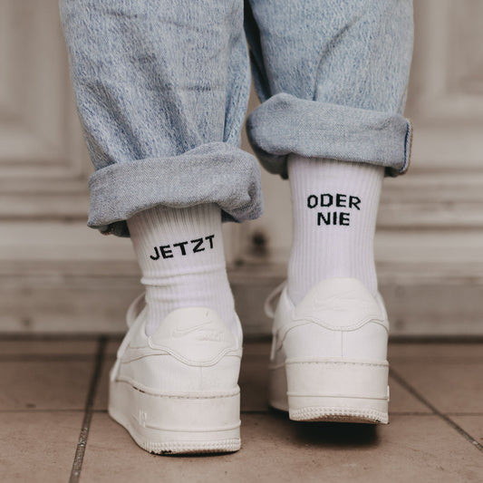Person in hochgekrempelter Jeans trägt weiße Socken mit schwarzem Schriftzug 'JETZT' auf der einen und 'ODER NIE' auf der anderen Ferse und weißen Sneakern mit Plateausohle.
