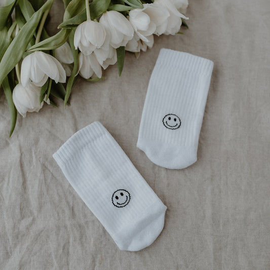 Zwei weiße sportliche Socken mit schwarzem Smiley, daneben ein Strauß weißer Tulpen auf Leinenstoff.