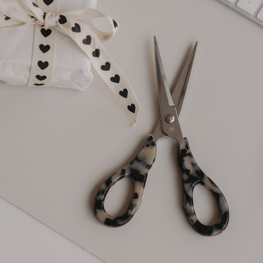 Silberne Schere mit Leo-Griff neben Geschenk mit Herzband auf hellem Hintergrund