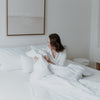 Eine junge Frau sitzt im Bett und guckt sich die Bettdecke an und die kleinen Herzen darauf. Hinter ihr sind noch 3 Kissen und über dem Bett hängt ein Bild.