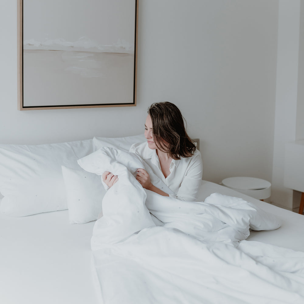 Eine junge Frau sitzt im Bett und guckt sich die Bettdecke an und die kleinen Herzen darauf. Hinter ihr sind noch 3 Kissen und über dem Bett hängt ein Bild.