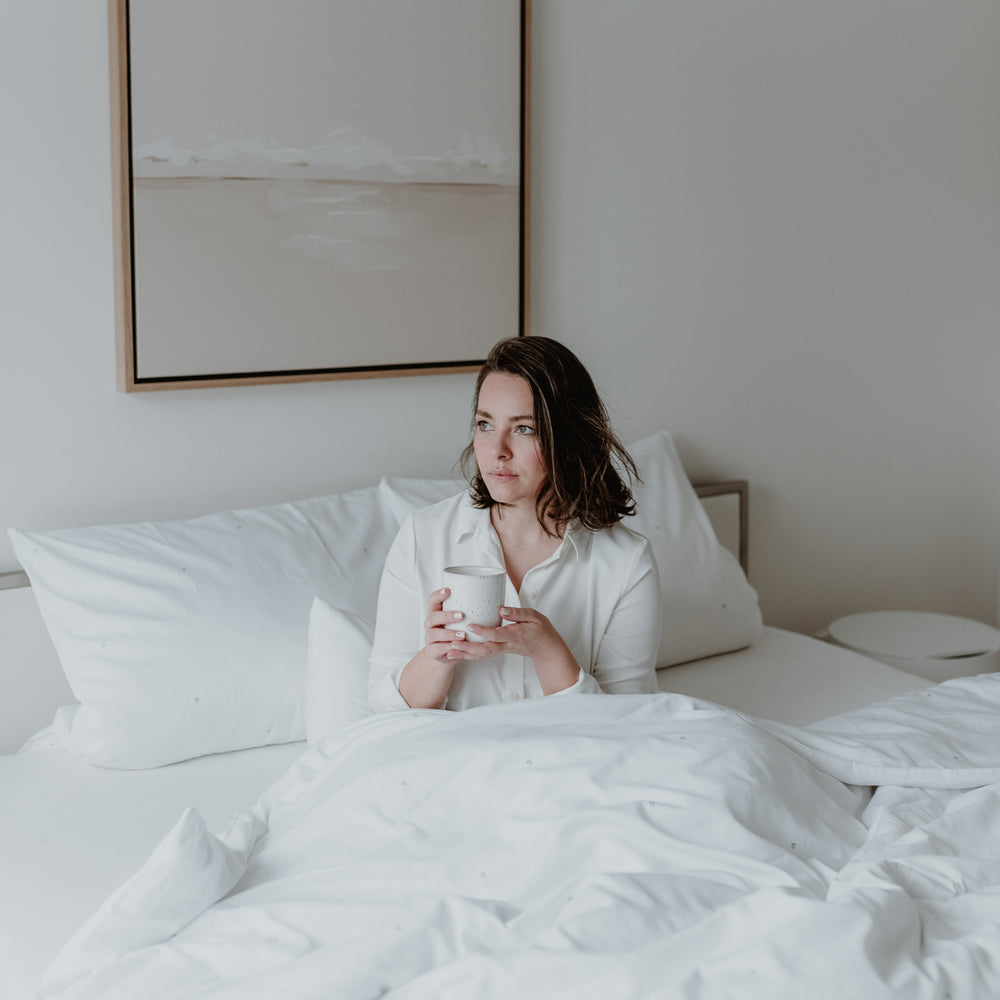 Eine junge Frau sitzt gemütlich mit einem Becher in ihrer Hand im Bett und freut sich über die gemütliche Bettwäsche.