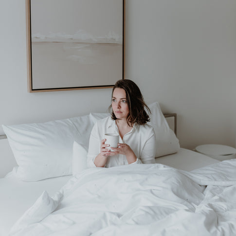 Eine junge Frau sitzt gemütlich mit einem Becher in ihrer Hand im Bett und freut sich über die gemütliche Bettwäsche.