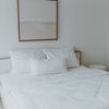 Ein wunderschön gemachtes Bett mit 2 größeren Kopfkissen und einem kleineren mittig davor haben alle kleine Herzen als Druck. Sehr minimalistisch, da es von weitem eher schlicht weiß erscheint. Über dem Bett hängt ein Bild. Auch eine Decke mit dem gleichen Druck ist zu sehen.