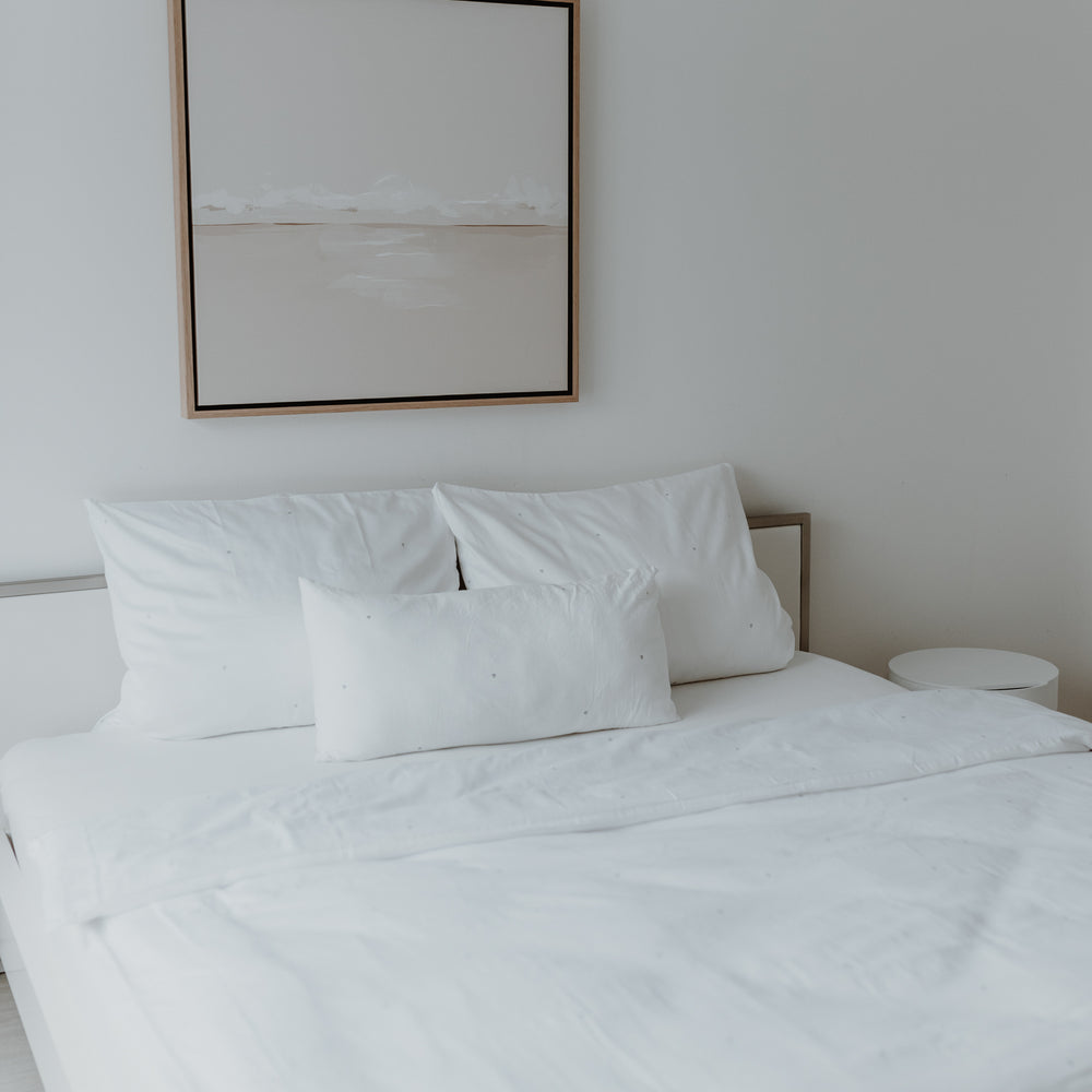 Ein wunderschön gemachtes Bett mit 2 größeren Kopfkissen und einem kleineren mittig davor haben alle kleine Herzen als Druck. Sehr minimalistisch, da es von weitem eher schlicht weiß erscheint. Über dem Bett hängt ein Bild. Auch eine Decke mit dem gleichen Druck ist zu sehen.