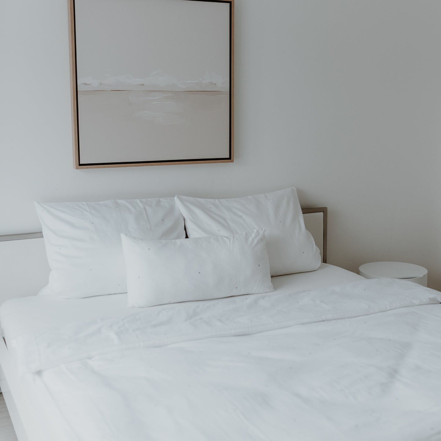 Ein wunderschön gemachtes Bett mit 2 größeren Kopfkissen und einem kleineren mittig davor haben alle kleine Herzen als Druck. Sehr minimalistisch, da es von weitem eher schlicht weiß erscheint. Über dem Bett hängt ein Bild. Auch eine Decke mit dem gleichen Druck ist zu sehen.