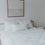 Ein wunderschön gemachtes Bett mit 2 größeren Kopfkissen und einem kleineren mittig davor haben alle kleine Herzen als Druck. Sehr minimalistisch, da es von weitem eher schlicht weiß erscheint. Über dem Bett hängt ein Bild. Auch eine Decke mit dem gleichen Druck ist zu sehen.