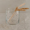 Glas mit schwarzen Punkten und getrockneten Ähren auf hellem Leinentuch im minimalistischen Stil fotografiert