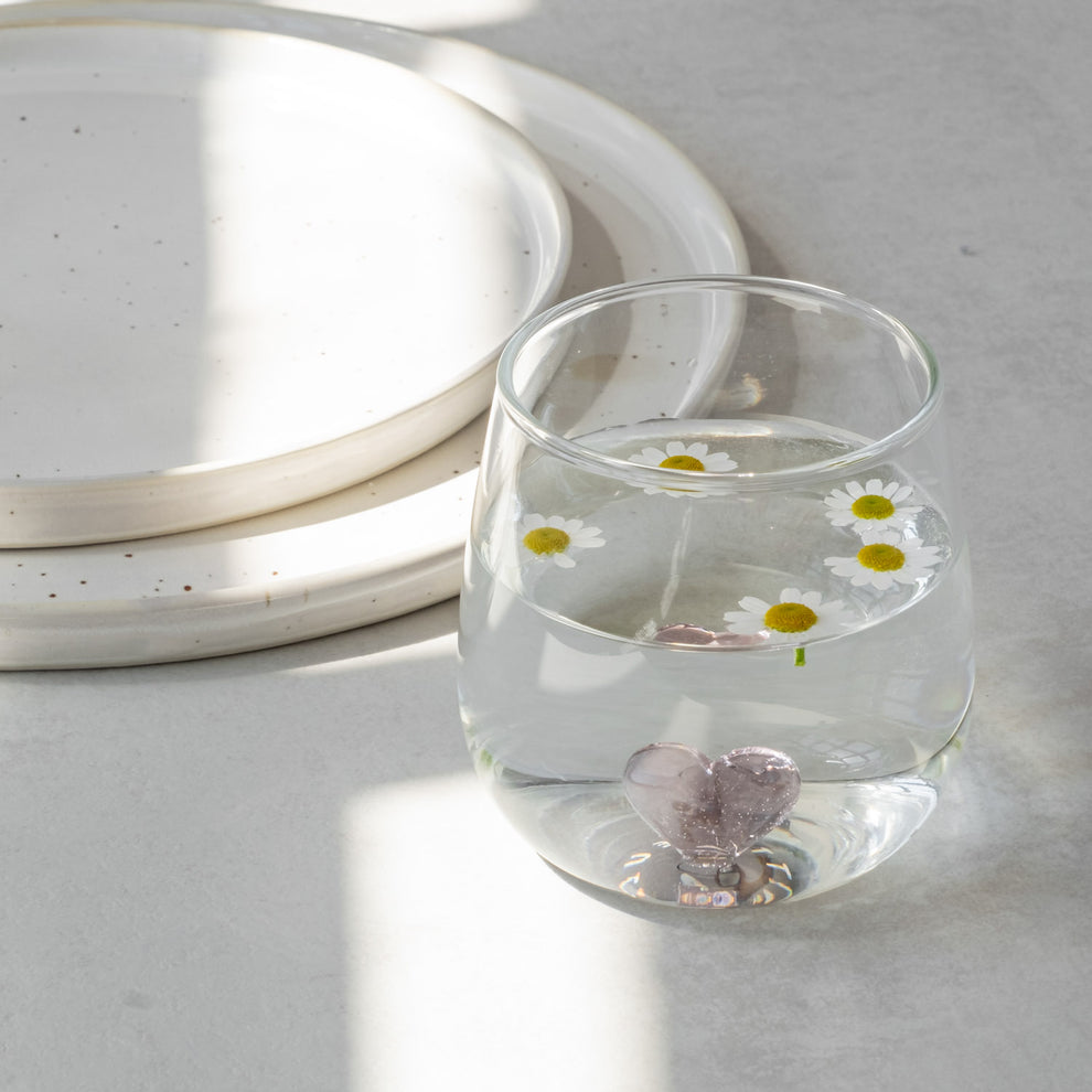 Trinkglas Herzfigur rosa mit Wasser und fünf kleinen Gänseblümchen, auf grauem Untergrund neben zwei Steingut Tellern.
