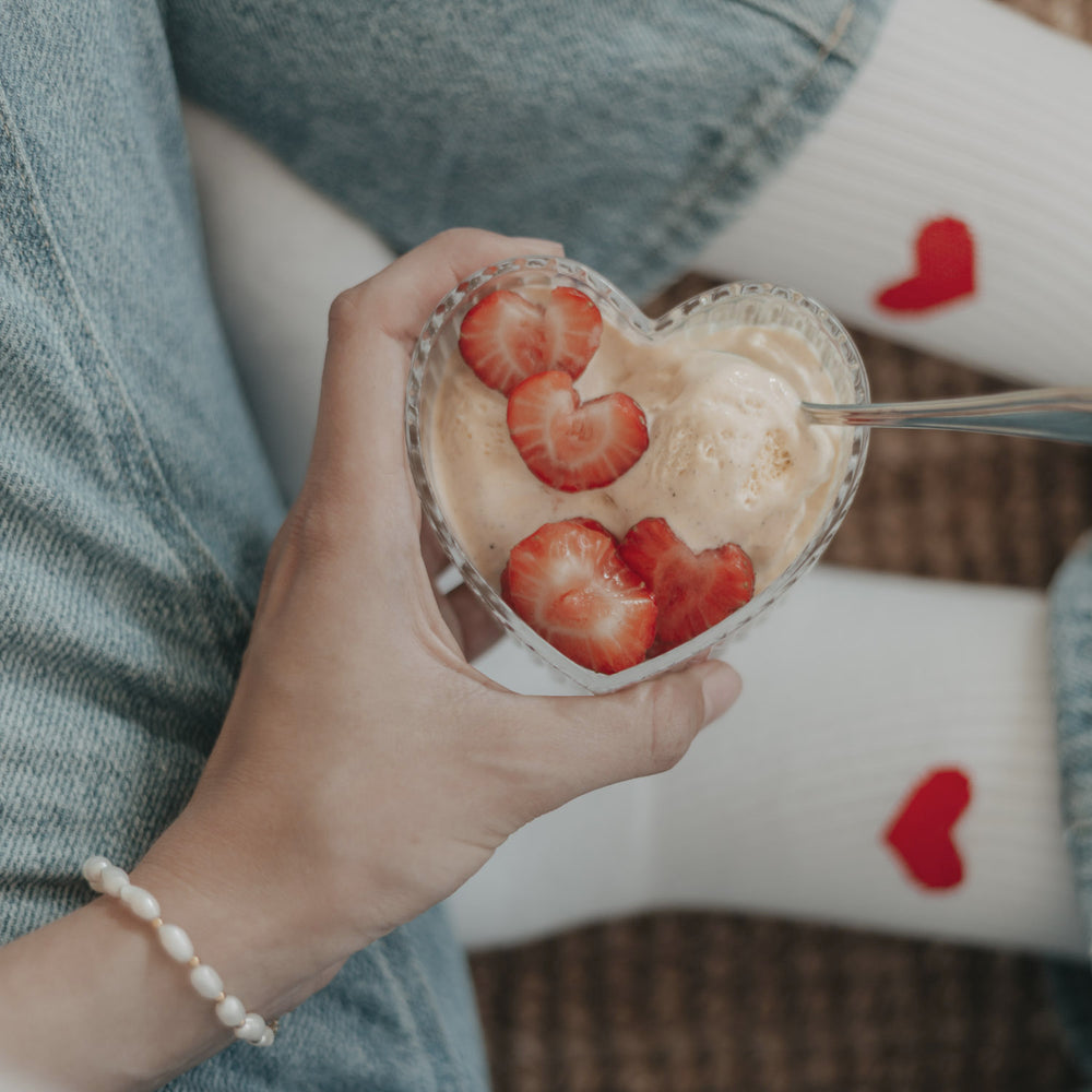 Hand hält eine Herzschale mit Vanilleeis und Erdbeeren, gemütlicher Snackmoment.