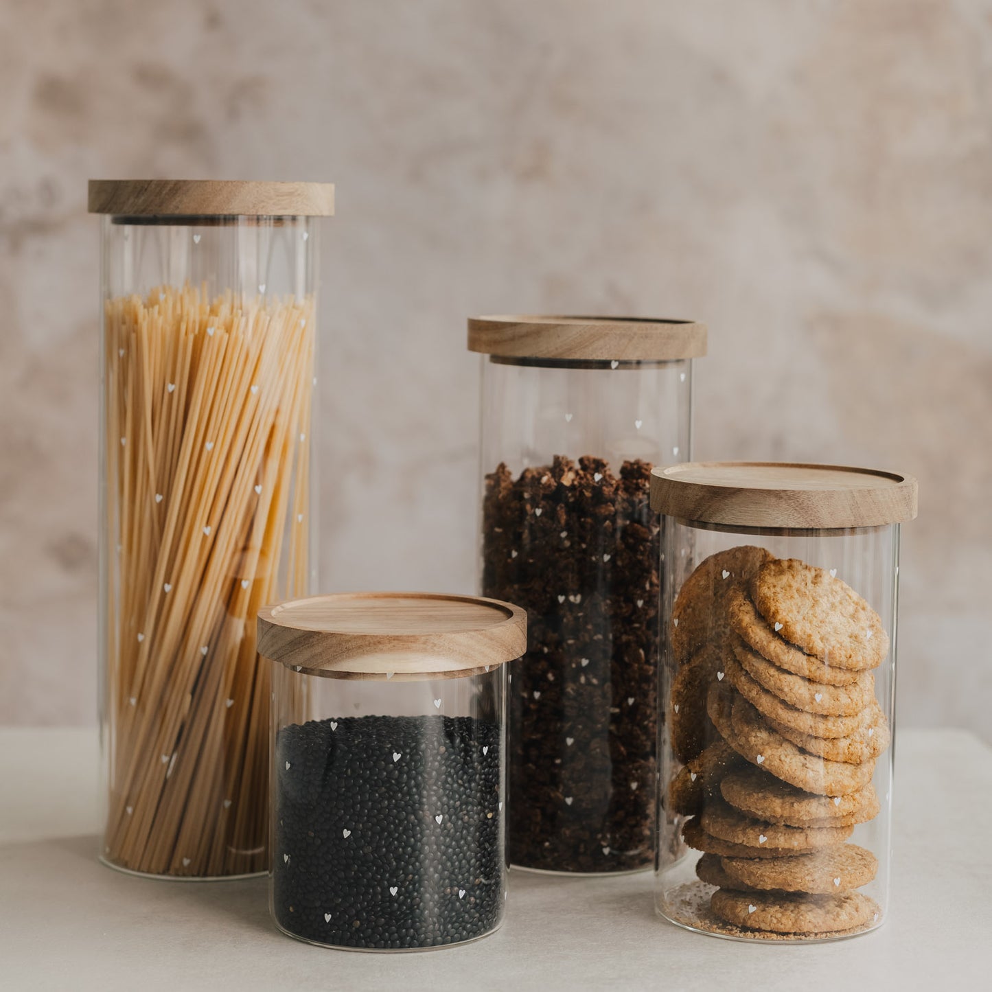Vier Vorratsgläser mit weißen Herzen und Holzdeckeln, gefüllt mit Spaghetti, Linsen, Müsli und Keksen