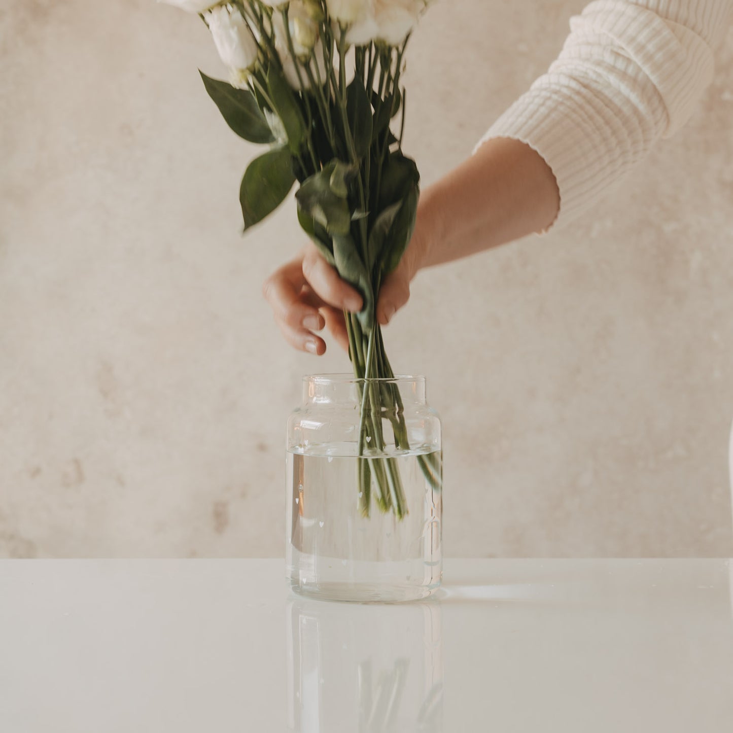 Hand stellt weißen Blumenstrauß in Glasvase mit Herzmotiv