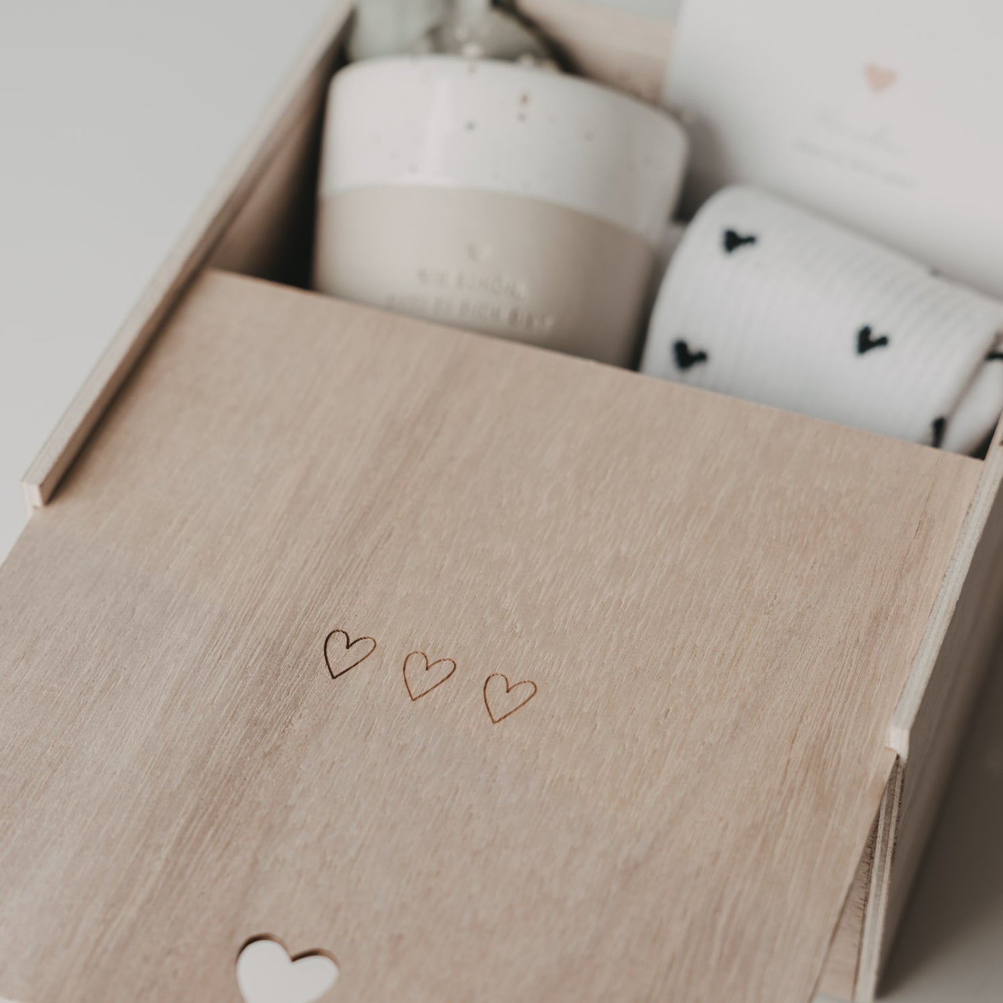 Detailaufnahme der halb geöffneten Erinnerungsbox mit drei eingravierten Herzen auf dem Holzdeckel. Im Inneren sind ein Keramikbecher, eine Karte und gemusterte Socken zu erkennen.