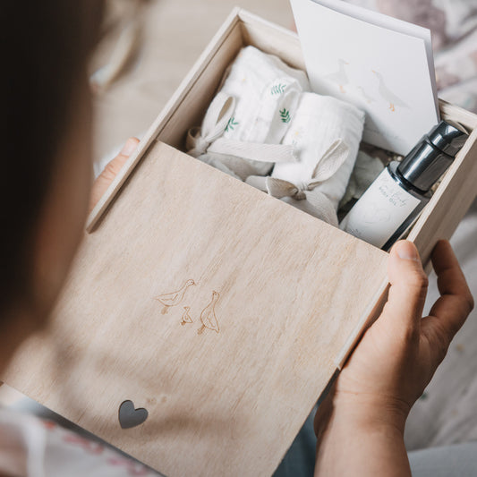 Person hält eine geöffnete Erinnerungsbox aus hellem Holz mit eingravierter Gänsefamilie in den Händen. Im Inneren befinden sich liebevoll arrangierte Musselintücher, eine Grußkarte mit Gänsemotiv und ein Pflegeöl für Mama und Baby.