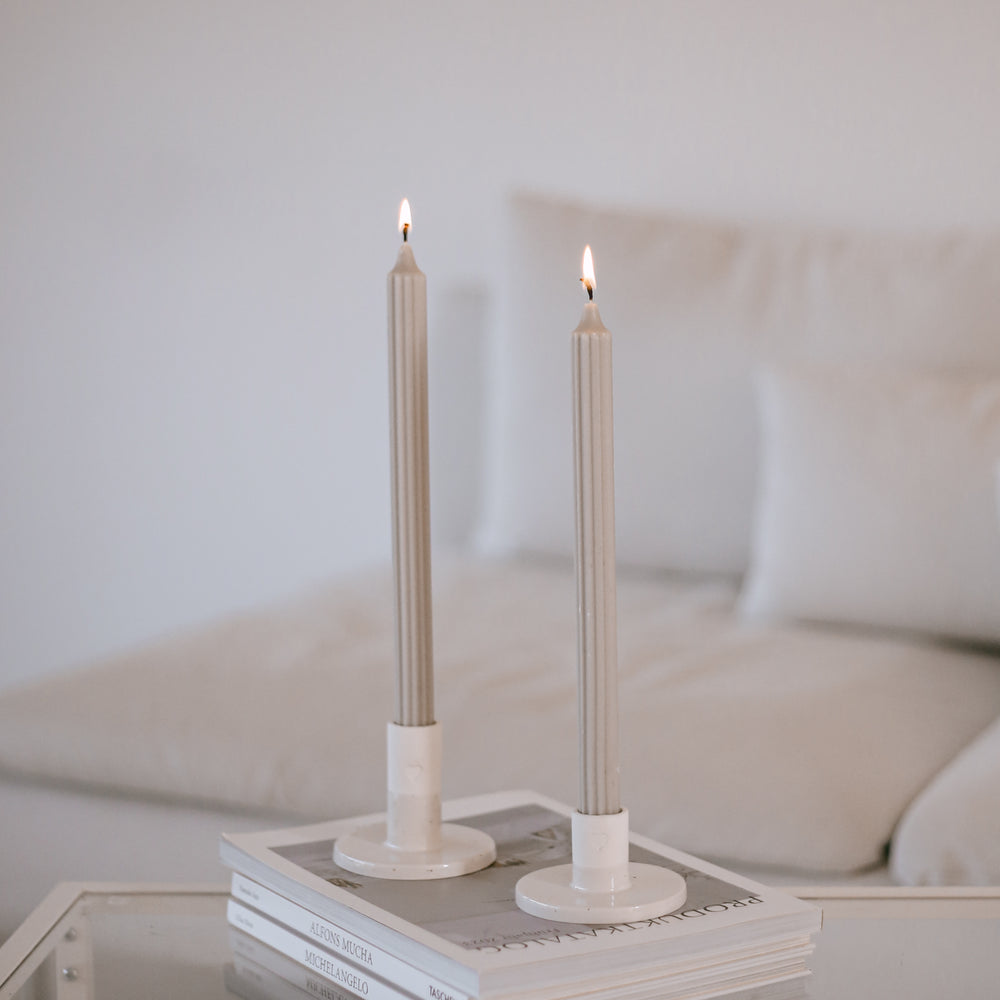 Zwei graue Stabkerzen in weißen Kerzenhaltern auf einem Glastisch vor heller Couch im Hintergrund
