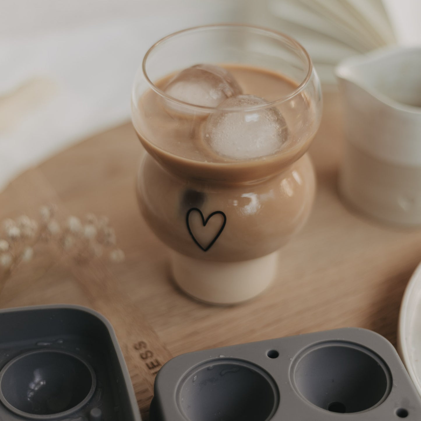 Eulenschnitt Bubble Glas mit schwarzem Herz-Print, gefüllt mit Iced Coffee und runden Eiswürfeln. Davor eine Silikoneiswürfelform auf einem runden Holzbrett.