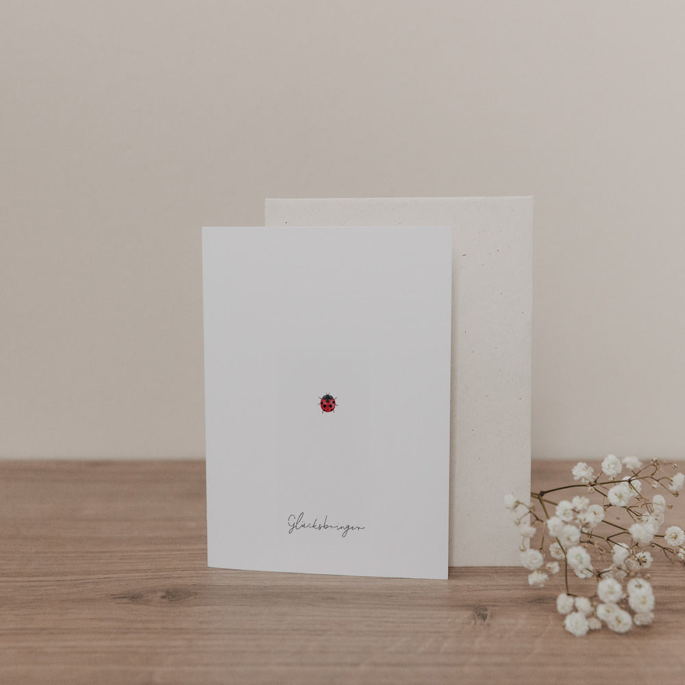 Minimalistische Klappkarte mit kleinem Marienkäfer-Motiv und Umschlag, liebevoll arrangiert auf Holzoberfläche mit weißen Blüten.