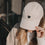 Beige Baseball Cap mit schwarzem Herz-Stick in Händen gehalten