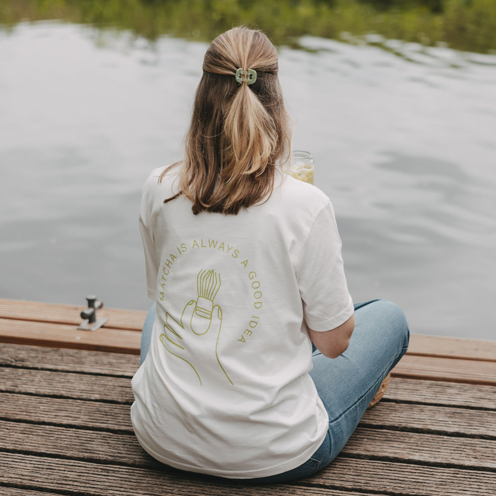 Frau mit weißem Matcha T-Shirt am See, Rückenansicht mit Getränk