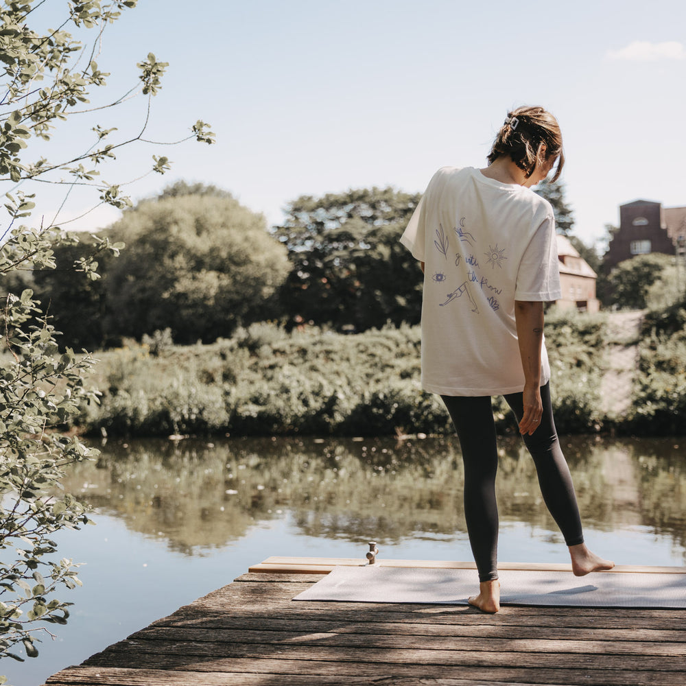 Frau auf Steg am See beim Yoga, lässiges weißes T-Shirt mit „go with the flow“ Rückendruck