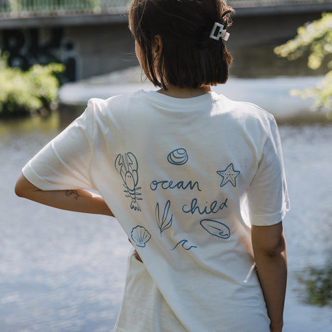 Rückenansicht weißes T-Shirt mit „Ocean Child“ Print und Meeresillustrationen am Wasser
