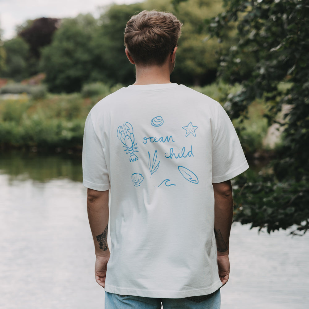 Rückenansicht Mann im weißen „Ocean Child“ T-Shirt mit blauem Print am Wasser