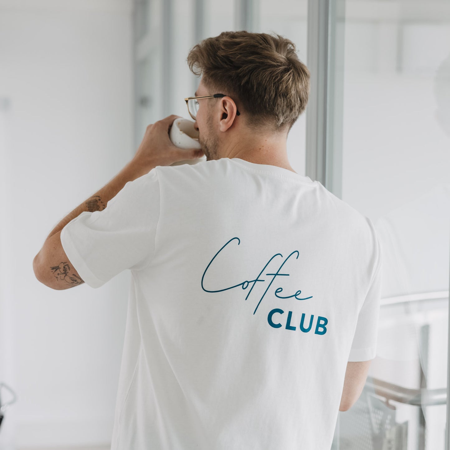 Mann im weißen „Coffee Club“ T-Shirt, Rückenansicht beim Kaffeetrinken