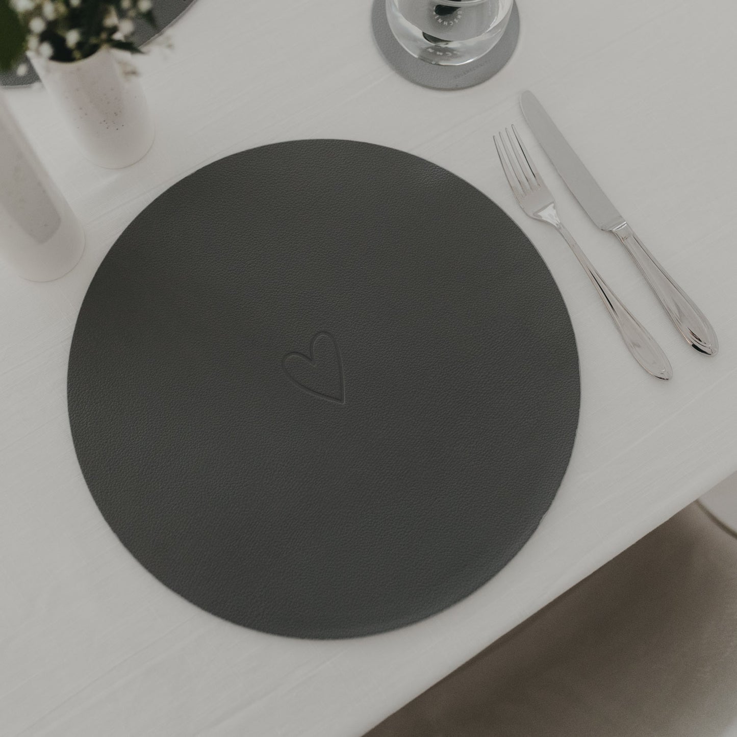 Dunkelgraues Platzset mit eingeprägtem Herz neben Besteck auf weiß gedecktem Tisch.