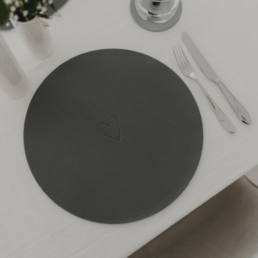 Dunkelgraues Platzset mit eingeprägtem Herz neben Besteck auf weiß gedecktem Tisch.