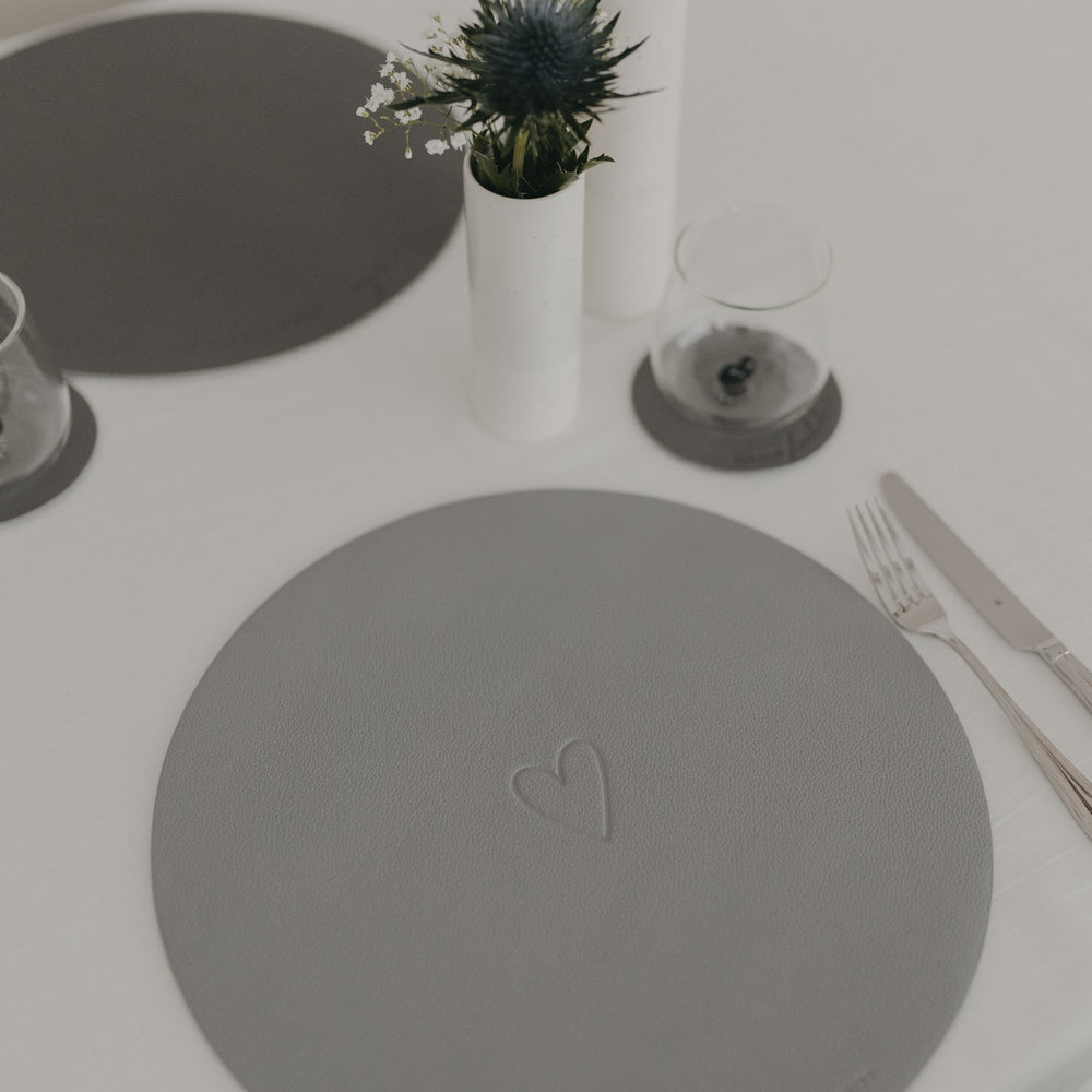 Helles graues Platzset mit Herzprägung neben Glas und Vase auf weißem Tisch.