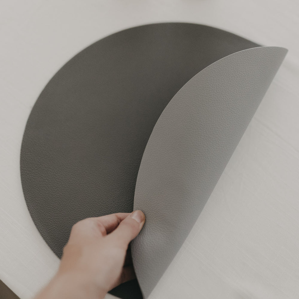 Hand dreht ein rundes, graues, wendbares Platzset mit strukturierter Oberfläche auf einem weißen Tisch um.