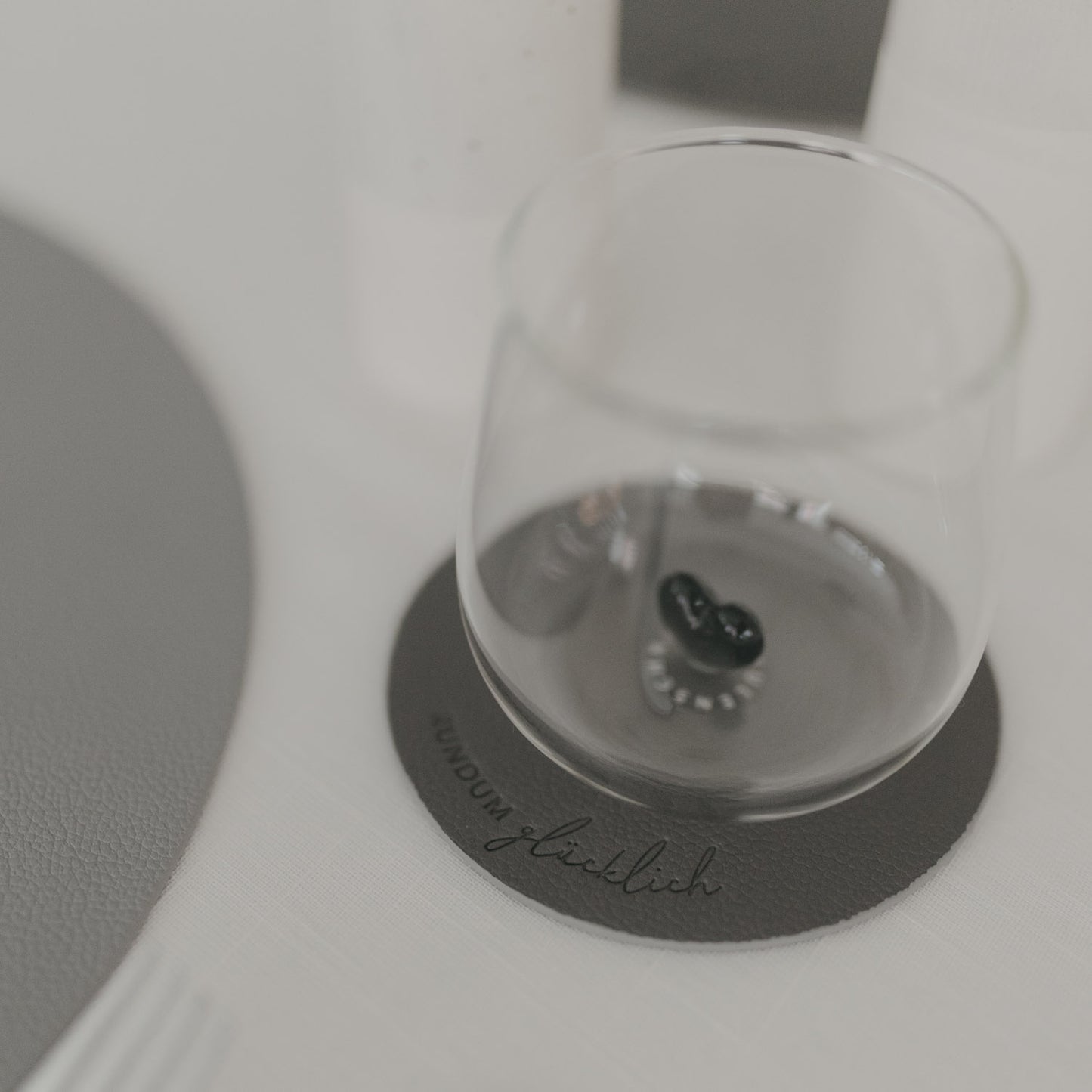 Grauer Untersetzer „Rundum glücklich“ mit Glas darauf, modernes Design auf hell gedecktem Tisch.