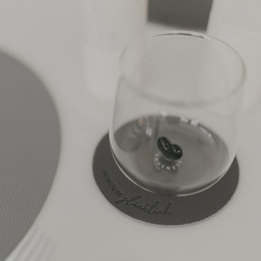 Grauer Untersetzer „Rundum glücklich“ mit Glas darauf, modernes Design auf hell gedecktem Tisch.