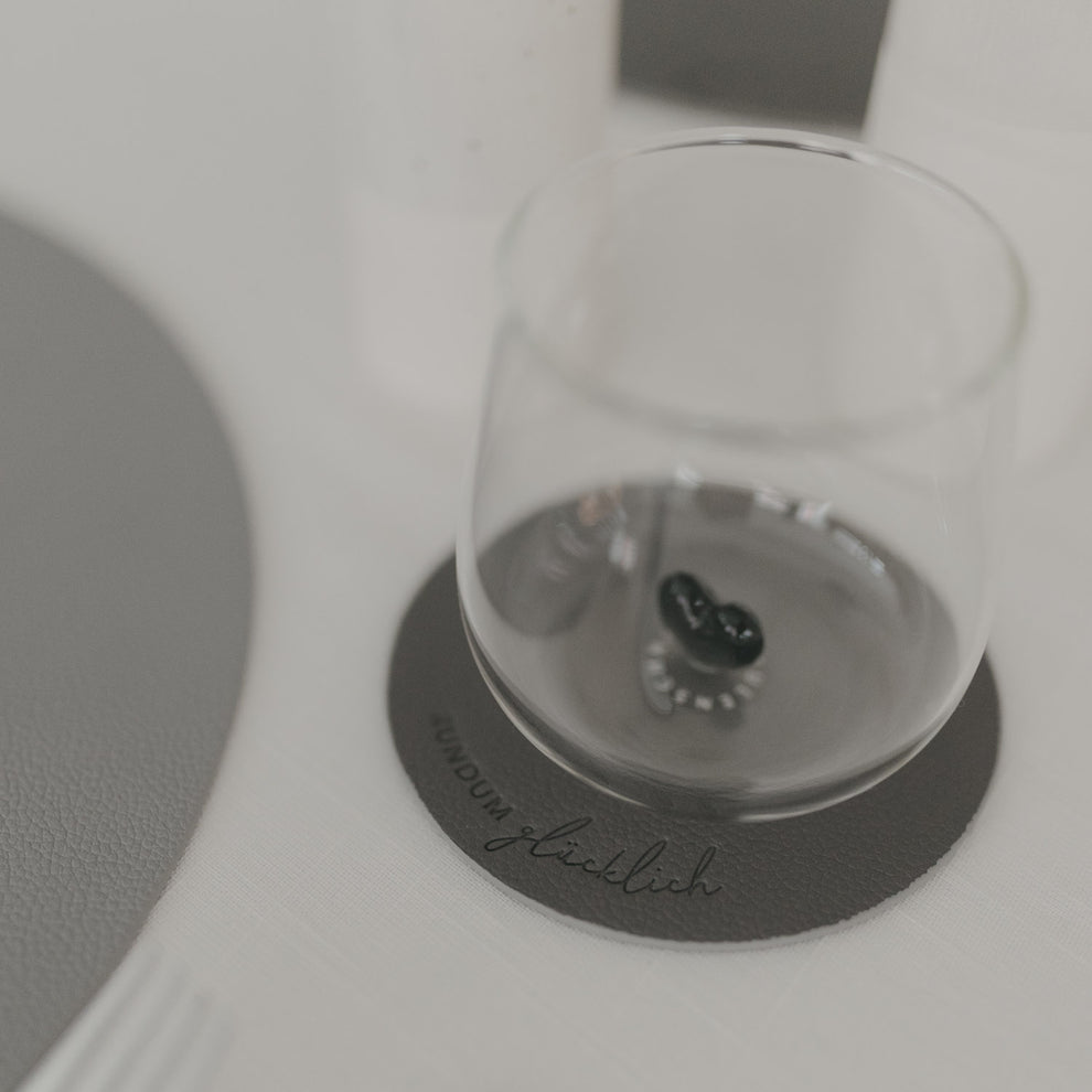 Grauer Untersetzer „Rundum glücklich“ mit Glas darauf, modernes Design auf hell gedecktem Tisch.