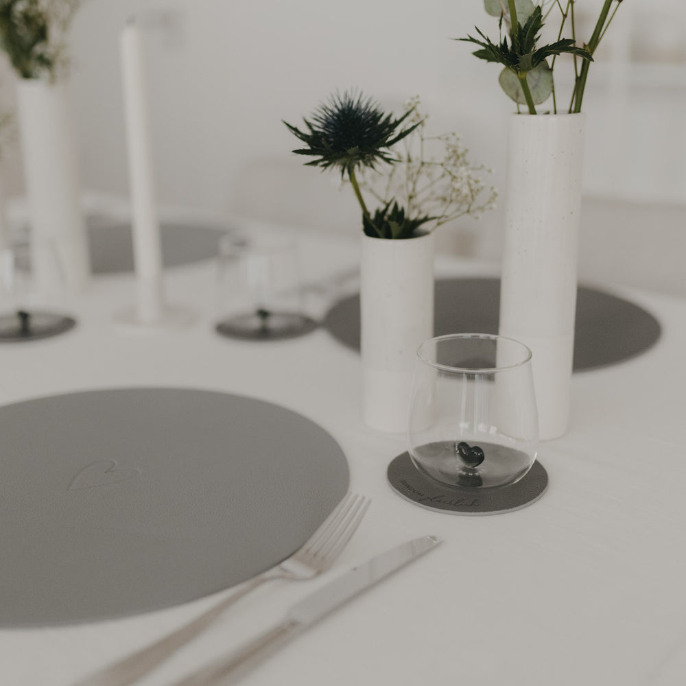 Grauer Untersetzer „Rundum glücklich“ auf elegant gedecktem Tisch mit weißen Vasen und grauen Platzsets.