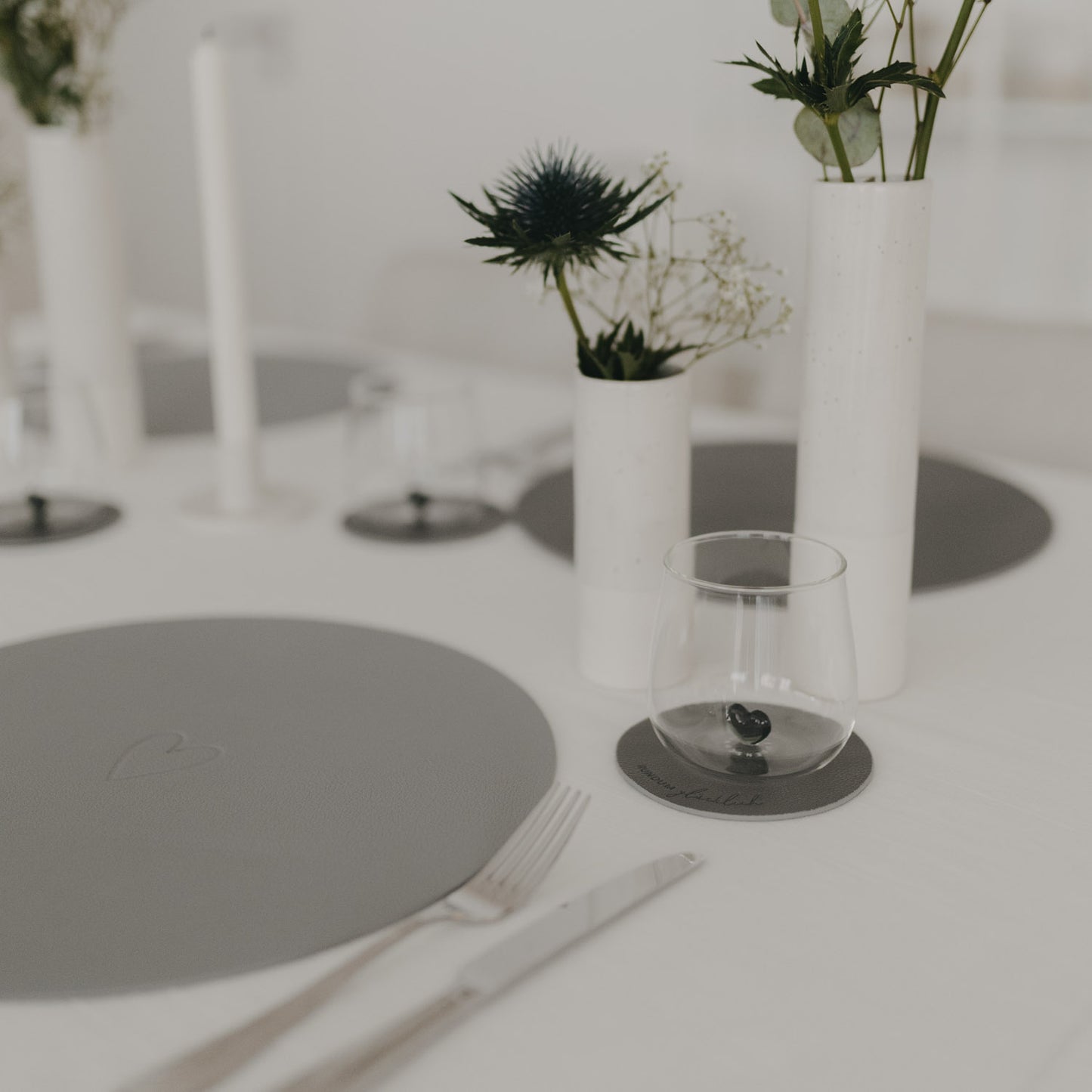 Grauer Untersetzer „Rundum glücklich“ auf elegant gedecktem Tisch mit weißen Vasen und grauen Platzsets.