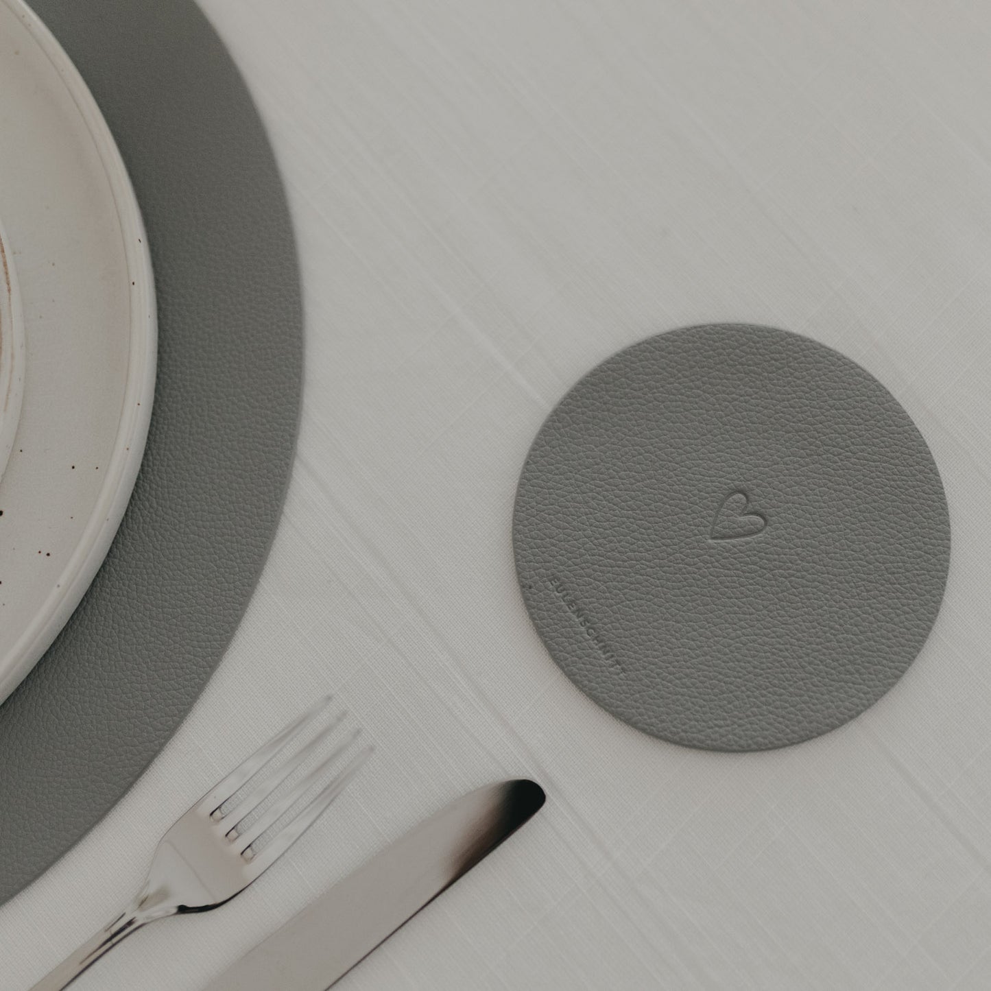 Dunkelgrauer Herz-Untersetzer neben Besteck und grauem Platzset auf weißer Tischdecke.