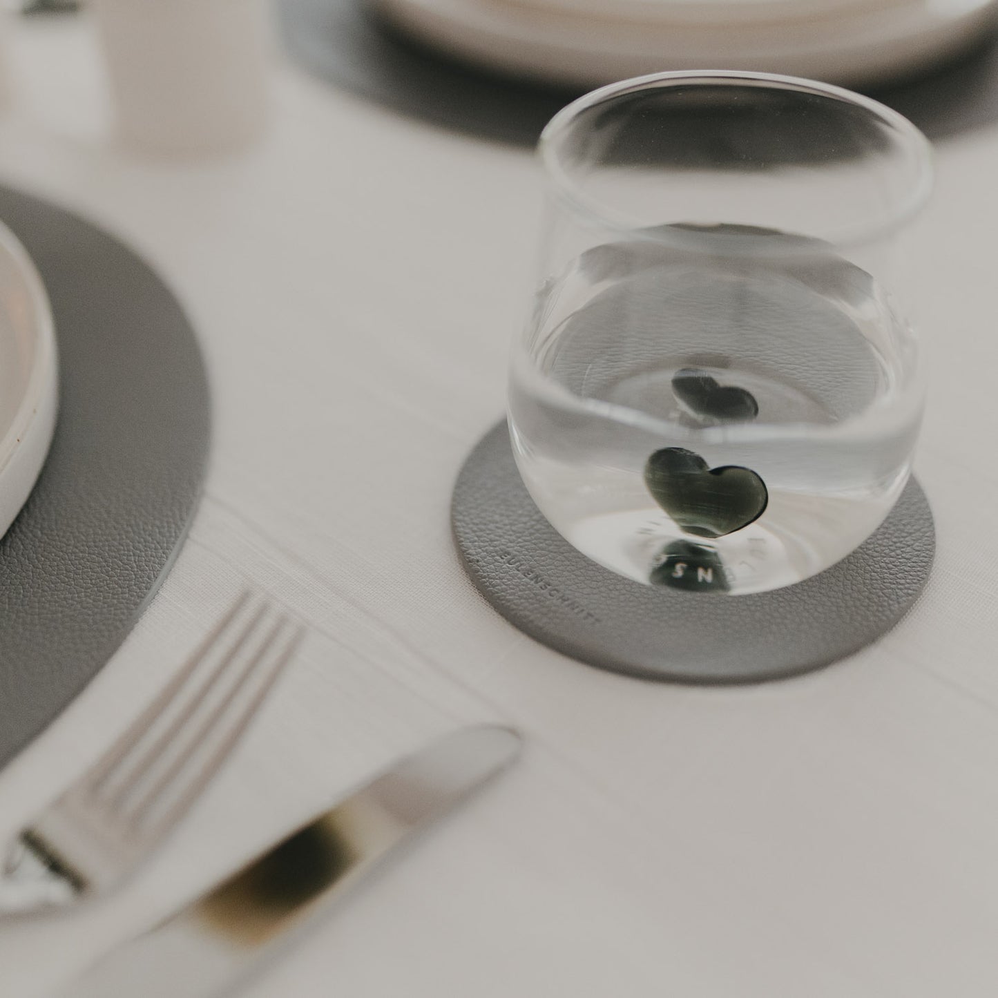 Glas auf grauem Untersetzer mit Eulenschnitt-Logo, passend zu grauem Platzset auf gedecktem Tisch.