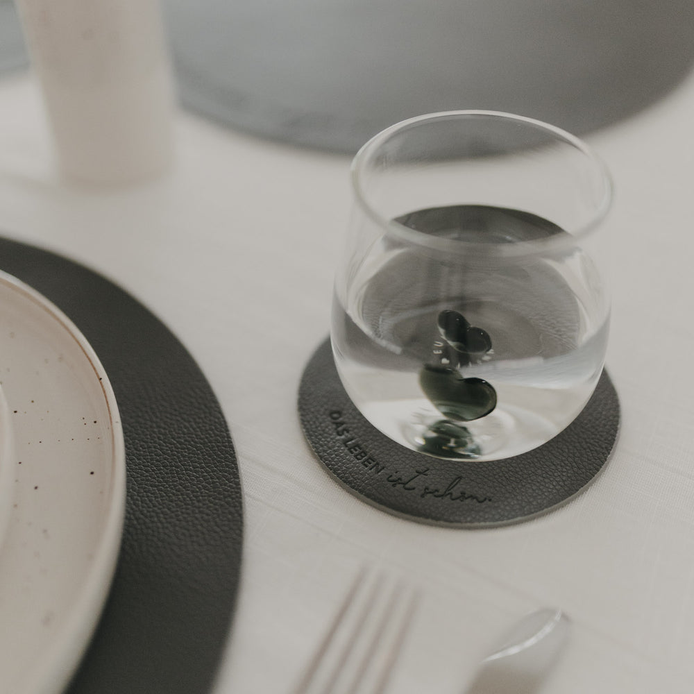 Glas auf dunkelgrauem Untersetzer mit Prägung ‚Das Leben ist schön‘ auf hell gedecktem Tisch.